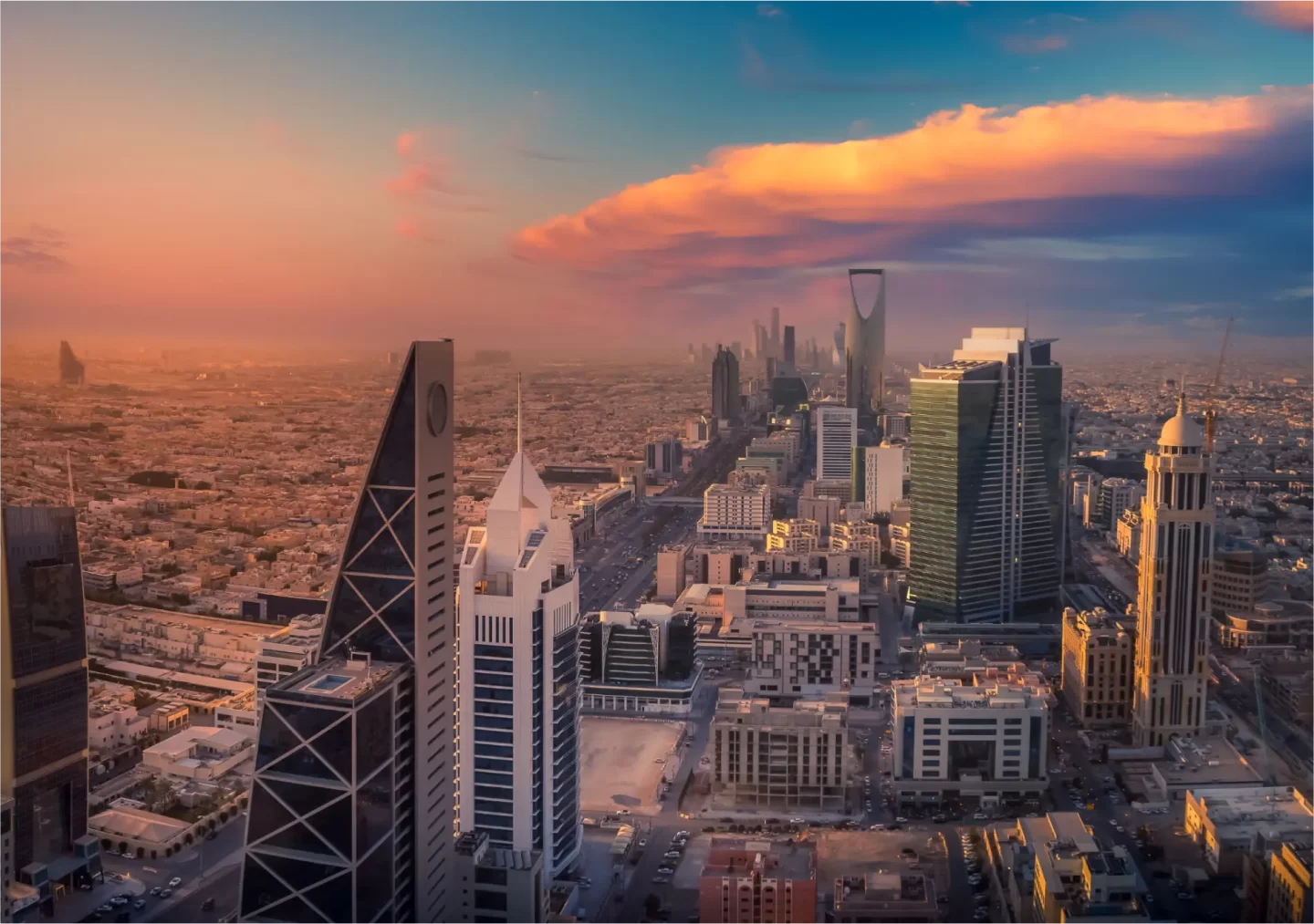 Aerial view of Riyadh's modern skyline at sunset, with prominent skyscrapers, including the Kingdom Tower, set against a colorful sky.